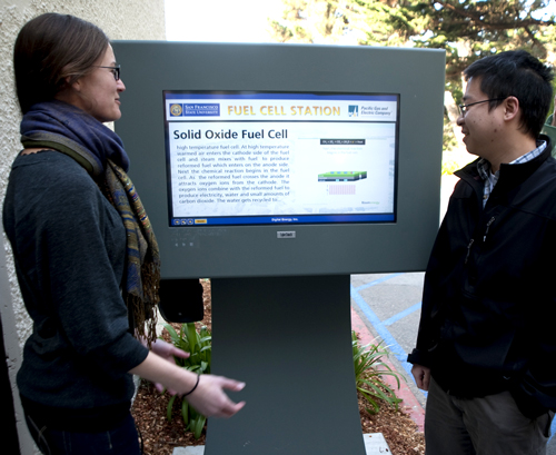 A photo of students from the Engineering Department demonstrating the educational touch-screen kiosk outside the fuel cell plant.