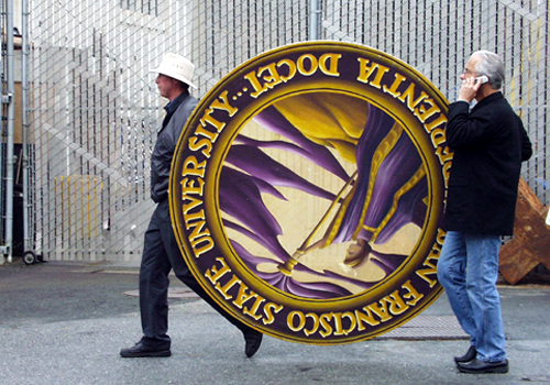 Photo of two staff members carrying a large wooden version of the University seal