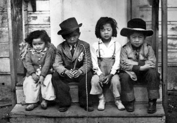 Black and white photo showing four young Filipino children sitting on a doorstep. One of them wears a kid-size McIntosh suit.