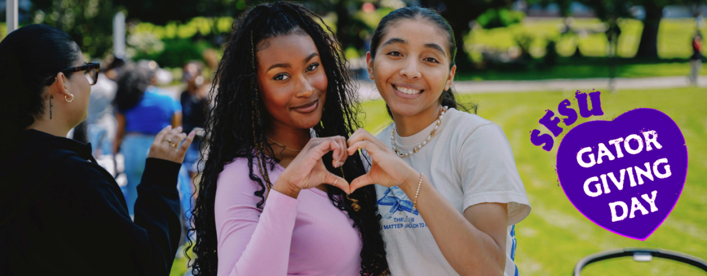 Two female students smiling and making a heart with their hands for Giving Day