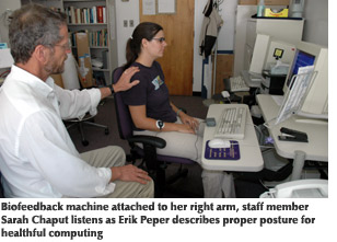 Photo of staff member Sarah Chaput with a biofeedback machine attached to her right arm listening to Erik Peper, director of holistic healing studies, describe proper posture for healthful computing