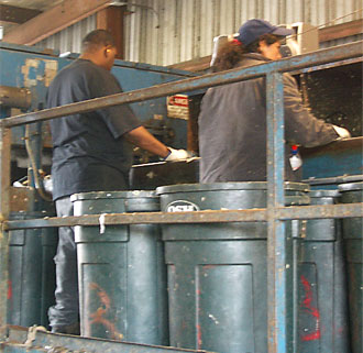 Photo of two green collar workers standing at a conveyor belt sorting recyclable materials at the Ecology Center in Berkeley. 
